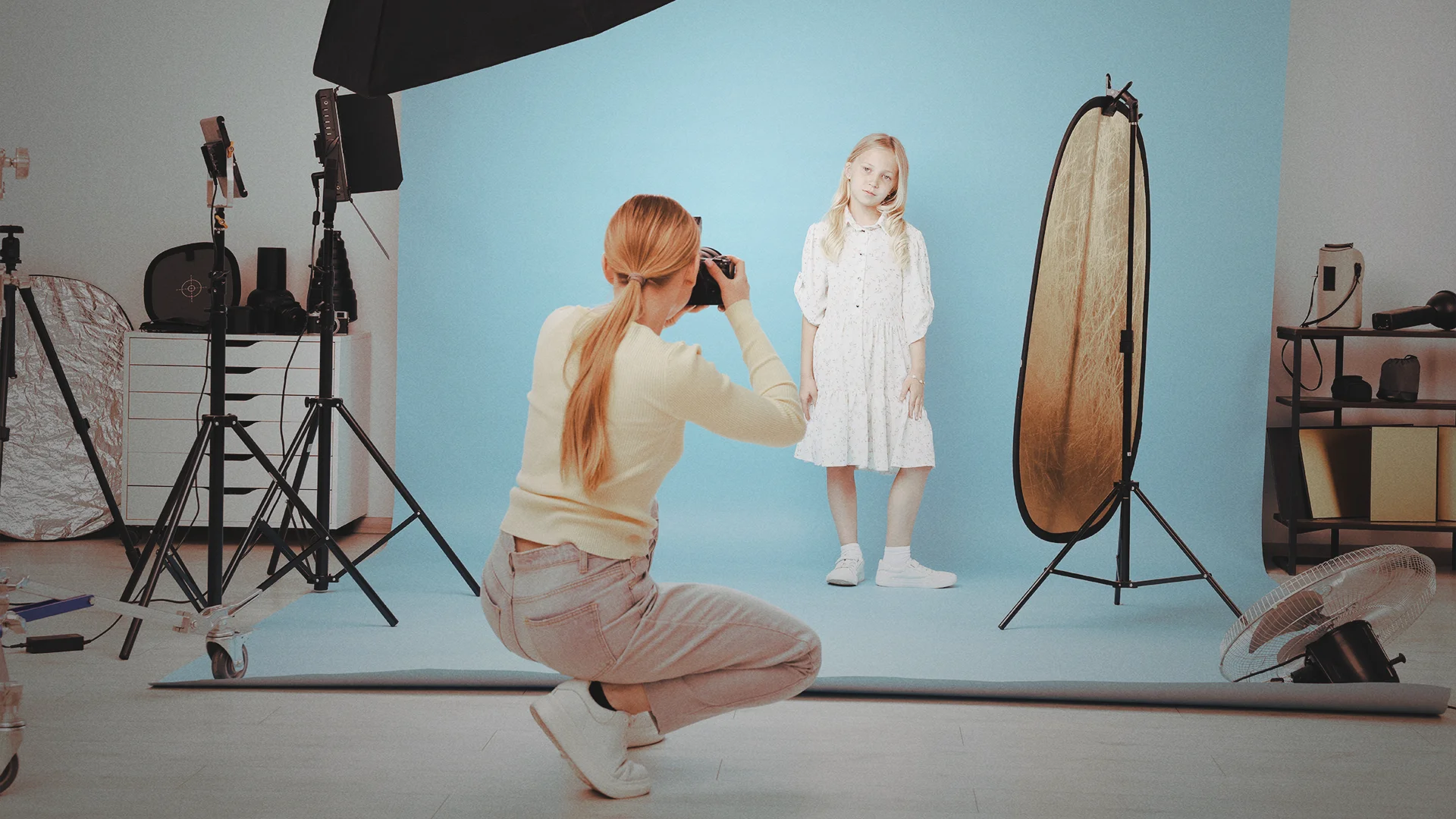 Im Vordergrund kniet eine Fotografin auf einem hellen Boden und hält eine Kamera vor ihr Gesicht. Sie trägt einen hellen Pullover, beige Jeans und weiße Sneaker. Ihre Haare sind zu einem niedrigen Pferdeschwanz gebunden. Vor ihr steht ein junges Mädchen mit langen blonden Haaren. Es trägt ein helles, knielanges Kleid und weiße Schuhe. Das Mädchen posiert vor einem großen, hellblauen Hintergrund, der nahtlos vom Boden zur Wand verläuft.